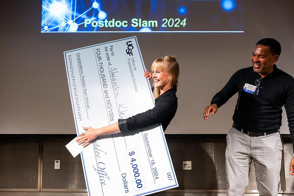 Alexandra Klein, Postdoc Slam 1st prize winner, holds a giant check as she celebrates her win